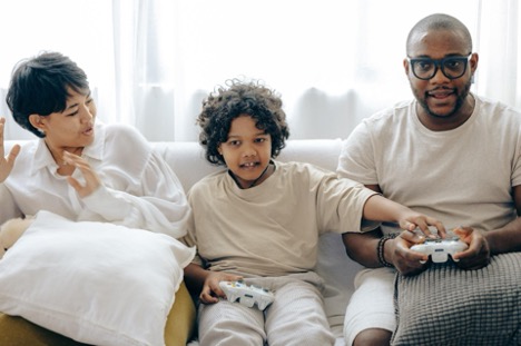 two adults sitting on a couch with a child in the middle. Child and adult male are playing a video game